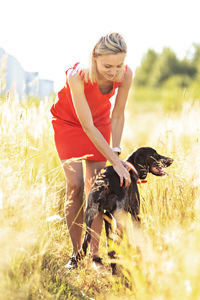 A young blonde woman in a bright dress stands in the middle of a field with a hunting dog.