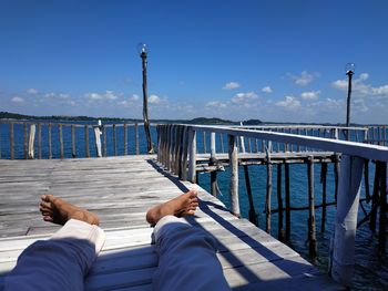 Low section of man relaxing on footbridge over sea against sky