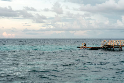 Couple sitting on jetty amidst sea against sky