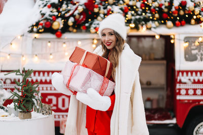 A beautiful woman with red lips holds gift boxes at a decorated christmas van on street in the city