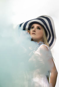 Thoughtful young woman wearing hat looking away by smoke against sky