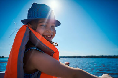 Portrait of cheerful girl wearing life jacket sitting against clear blue sky