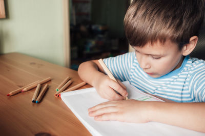 Cute boy sits at desk and draws in album with colored pencils
