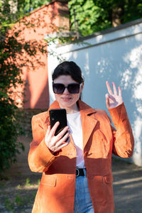 A young brunette girl in an orange jacket takes a selfie against a brick wall