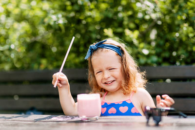 A cute baby is drinking a berry smoothie through a straw, sitting on a bench in the garden. 