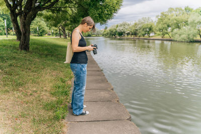 Woman 40s reviewing photos on camera by urban riverbank with tote bag