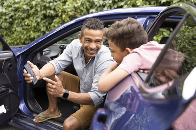 Excited father showing mobile phone to son leaning on car door