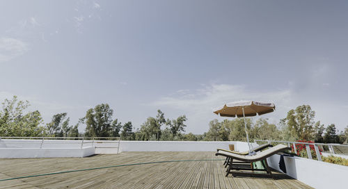 Two sunbeds in the sun and sunshade on the wooden dais with the blue sky and trees in the background