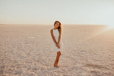 Full length of young woman standing at beach against sky during sunset