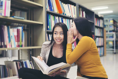 Young woman whispering to female friend studying in library