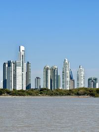 Buildings by river against clear blue sky