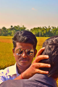 Man wearing sunglasses touching friend at field against clear sky