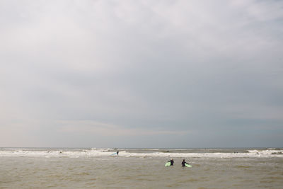 Distant view of people with surfboards in sea against cloudy sky