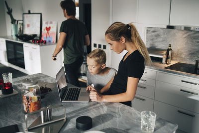 High angle view of boy standing by sister using laptop at kitchen island