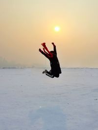 Person jumping over snow covered field against sky during sunset