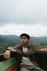 Young man sitting on car trunk against sky