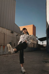 Full length of stylish self assured young ethnic female millennial in trendy outfit looking at camera while standing in urban area against cloudless blue sky with raised leg