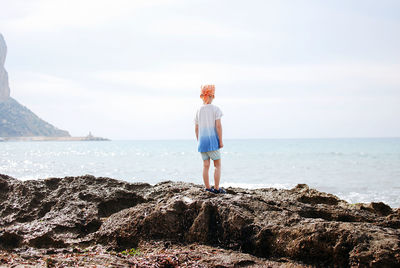 Rear view of little boy in orange bandana standing on rock at beach against sky