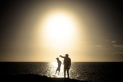 Silhouette man playing with dog at beach against sky during sunset