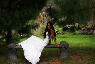 Portrait of young woman wearing white dress while relaxing on seat at park