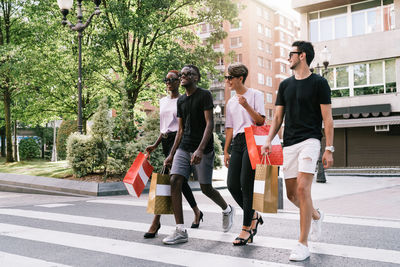 Full length of cheerful young diverse students in trendy outfits and sunglasses carrying purchases in shopping bags and chatting happily while crossing road on city street