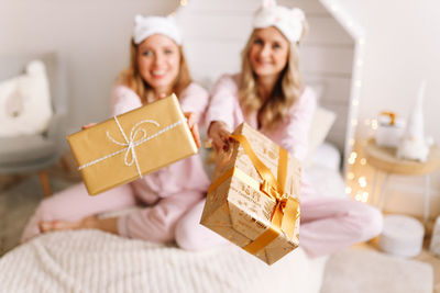 Two happy smiling girls hold christmas gift boxes in their hands while sitting on the bed at home