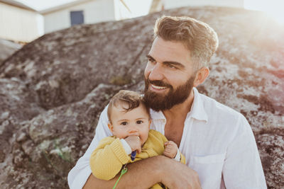 Portrait of baby girl with father looking away while sitting on rock formation