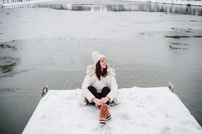 Happy woman with eyes closed sitting on pier in front of frozen lake in city