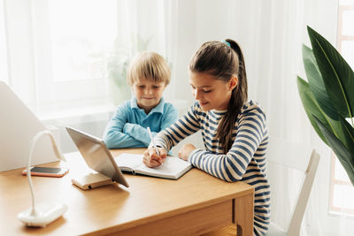 A schoolgirl, sitting at a table with her younger brother, performs an assignment on a tablet 