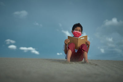 Full length of man reading book at beach against sky