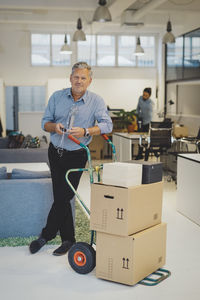 Full length portrait of confident mature businessman standing by luggage cart with stacked cardboard boxes in new office