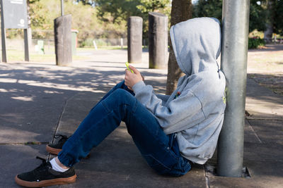 Side view of teenage boy using phone while sitting on footpath