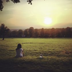 Rear view of woman with dog sitting on grassy field against sky during sunset