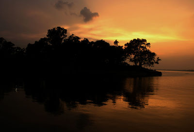 Silhouette trees by lake against orange sky