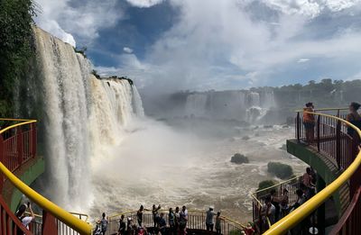 People looking at waterfall against cloudy sky