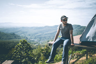 Young man sitting on mountain against sky