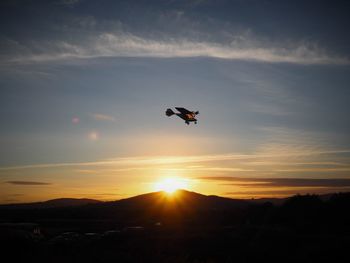 Low angle view of light aircraft flying over silhouette mountains during sunset