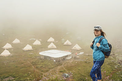 Young woman in cap blue with backpack hiking in mountain valley against tent camping in fog view