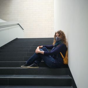 Young curly woman in a protective mask and jeans sits on the stairs of an empty museum building