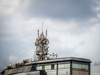 Low angle view of communications tower on building roof against cloudy sky