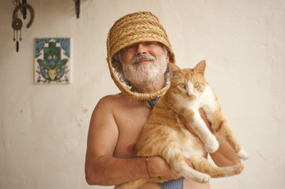 Portrait of shirtless man with basket on head holding cat while standing against wall