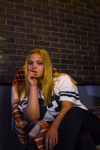 Portrait of young woman sitting against brick wall at home