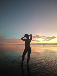 Silhouette young woman standing at beach against sky during sunset