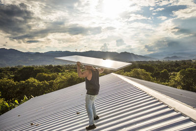 Construction worker carries solar panel above head on roof.