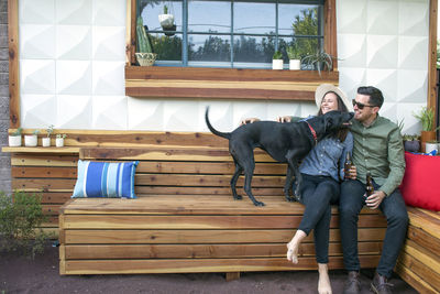 Happy couple holding beer bottles playing with dog while sitting in backyard