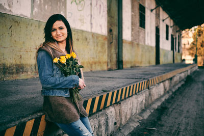 Portrait of smiling young woman holding roses while leaning on retaining wall