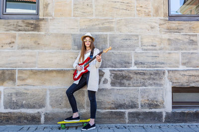 Portrait of young woman playing guitar while standing with skateboard by wall