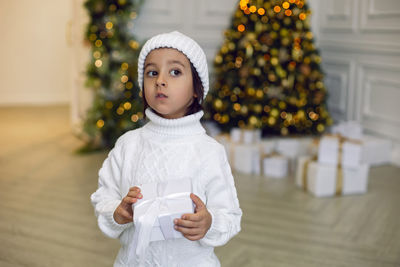 Boy in a white knitted sweater and hat stands with gift box  christmas tree at home on christmas day