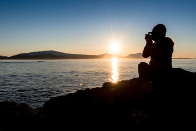 Silhouette woman photographing at beach against clear sky during sunset