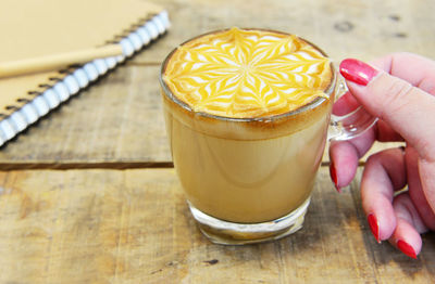 Women holding frothy coffee by book and pen kept on wooden table